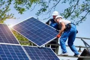 Two young technicians mounting heavy solar photo voltaic panel on tall steel platform on green tree background. Exterior solar panel voltaic system installation, dangerous job concept.